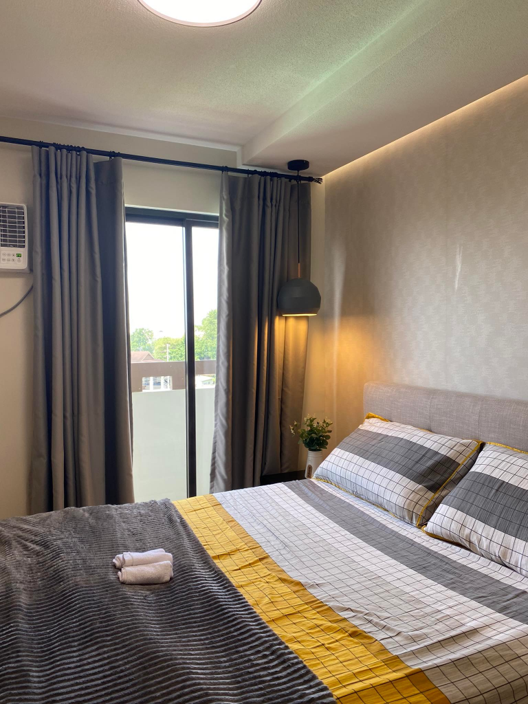 The bedroom of the 1-bedroom unit, featuring a comfortable bed with stylish bedding, floor-to-ceiling curtains, and a modern pendant light. The image emphasizes the unit's relaxing and well-designed sleeping area.