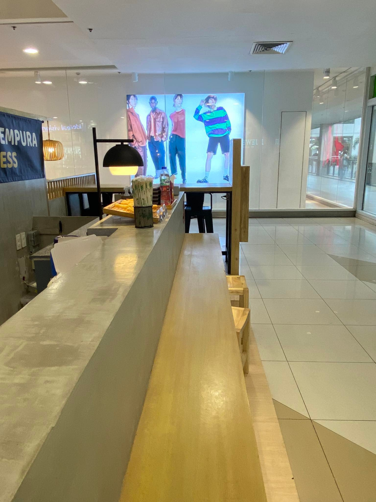 The modern design of Mr. Tempura Express is further emphasized by this image of its sleek counter and simple wooden seating area within a shopping mall.