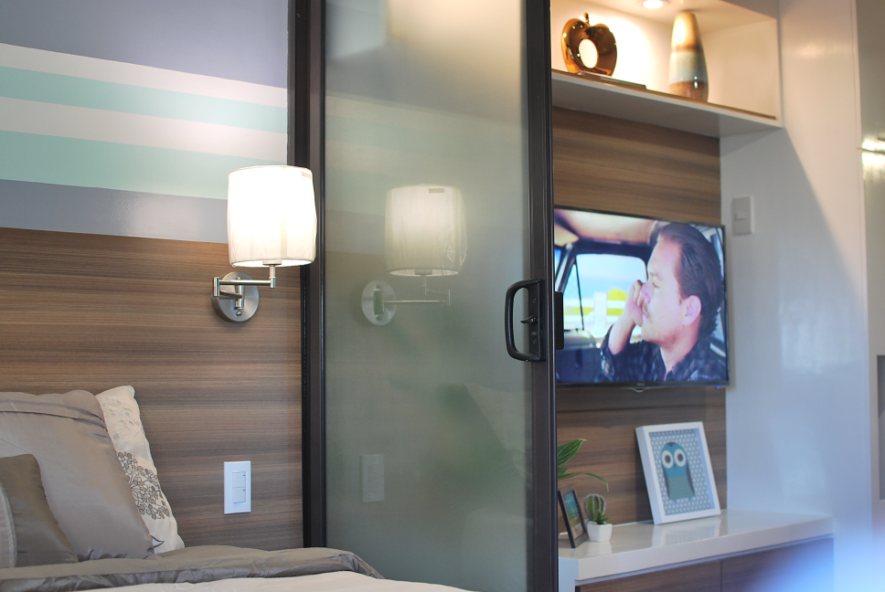 The bedroom area of a 1-bedroom unit, showcasing a stylish design with built-in shelving, a wall-mounted TV, and a frosted glass sliding door for separation.