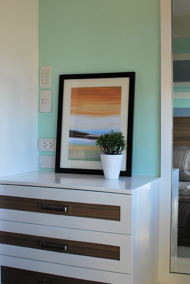 A detail shot showcasing the stylish wall decor and lighting in the unit, featuring a framed artwork and a small potted plant on a white surface. The image highlights the unit's calm and sophisticated aesthetic.