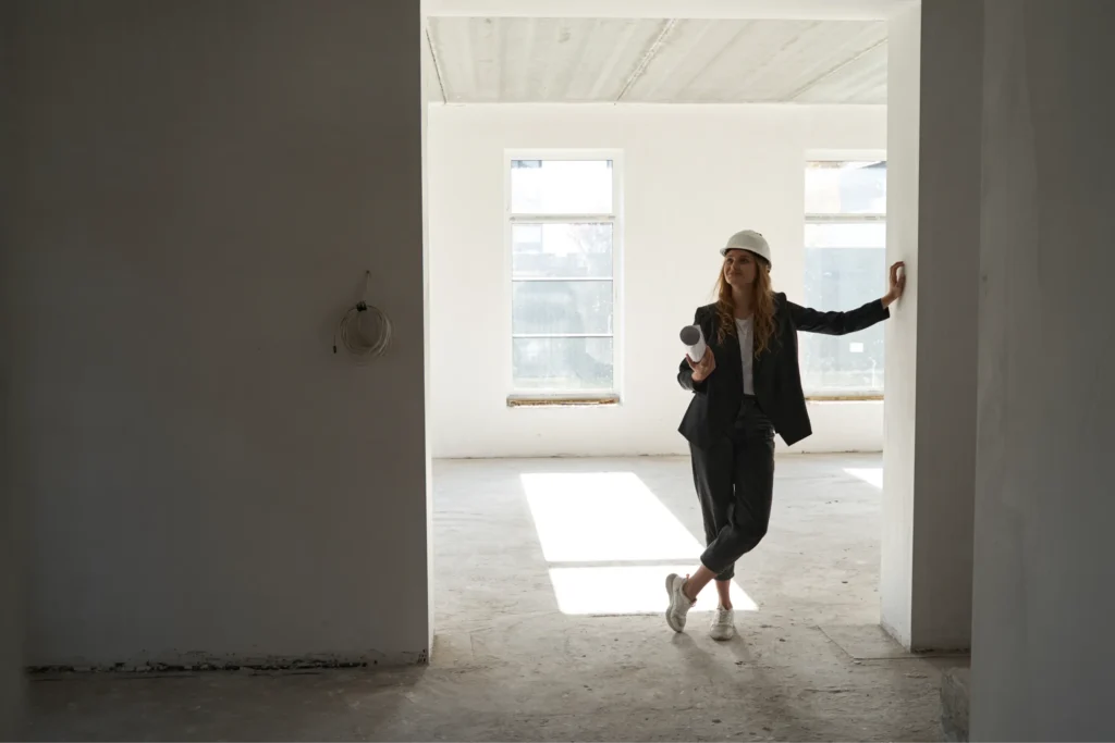 An interior designer, wearing a hard hat, stands in an empty room, reviewing blueprints. The image conveys the importance of on-site assessment in the interior design process.